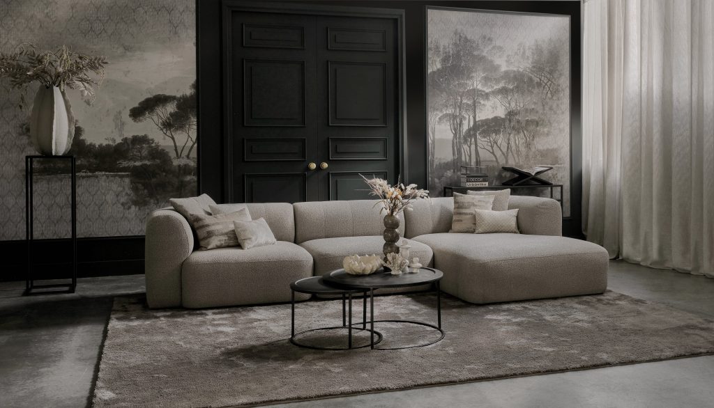 Elementenbank Emanuelle met een longchair. Uitgevoerd in een lichte beige stof. Gecombineerd met een groot vloerkleed en naturel decoratie.